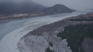 AK0001_1300 - 4K aerial stock footage flying over icy river toward Glenn Highway bridge, Knik River Valley, Alaska in snow