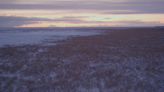 AK0001_1701 - 4K aerial stock footage flying low over frozen, snowy ground and grass at twilight, Point MacKenzie, Alaska