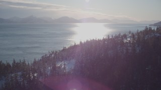 AK0001_1837 - 4K aerial stock footage fly over snowy, wooded shore, sun reflecting off water, Port Wells, Alaska