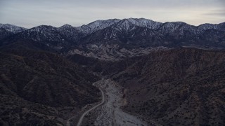 AX0008_003 - 5K aerial stock footage fly over a dry riverbed toward rock formations and snowy mountains at twilight, San Gabriel Mountains, California