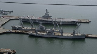 AX0175_0217 - 6K aerial stock footage of passing an aircraft carrier and Naval ships docked in Alameda, California