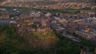 AX112_080E - 5.5K aerial stock footage of Edinburgh Castle on a hill with a view of cityscape, Scotland at sunset
