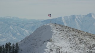 AX125_210 - 5.5K aerial stock footage approach American flag waving atop a snowy peak in winter, Oquirrh Mountains, Utah