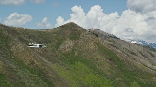 AX140_139E - 5.5K aerial stock footage of a Tecnam P2006T passing a green mountain, aspen trees, snow patches, Wasatch Range, Utah