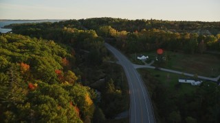 AX149_124E - 5.5K aerial stock footage flying over forest road with light traffic, fall foliage, in autumn, Stockton Springs, Maine, sunset