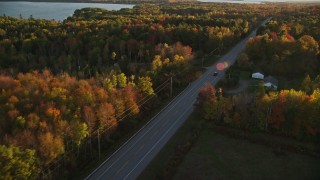AX149_130E - 5.5K aerial stock footage tracking car on road through forest in autumn, Stockton Springs, Maine, sunset