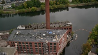 AX152_131 - 5.5K aerial stock footage orbiting away from an abandoned factory and smoke stack along the river, autumn, Lowell, Massachusetts