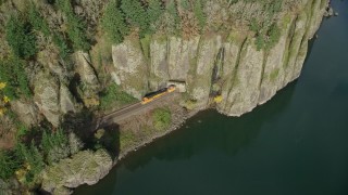 AX154_012E - 5.5K aerial stock footage revealing train tracks and a train emerging from the cliffs of Columbia River Gorge, Washington