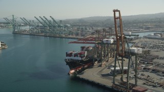 AX68_170 - 4.8K aerial stock footage of passing a cargo ship with containers beneath cranes at the Port of Los Angeles, California