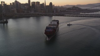 DFKSF10_052 - 5K aerial stock footage of a cargo ship sailing under Bay Bridge, reveal Downtown San Francisco skyline, California, sunset