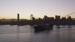 DFKSF10_053 - 5K aerial stock footage of a cargo ship sailing under Bay Bridge near Downtown San Francisco skyline, California, sunset